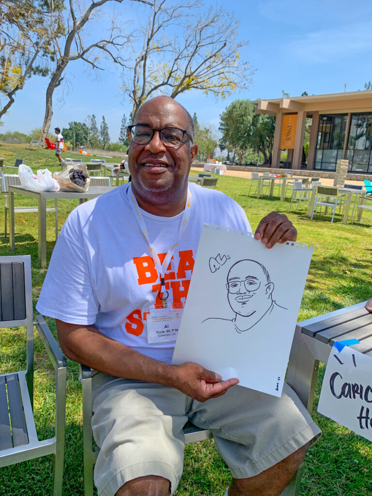 An alum holds up a cartoon drawing of himself while sitting on the mounds. 