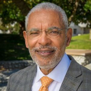 Melvin Oliver has close-shaved gray hair and beard with a gray dress jacket over a white collared shirt with an orange tie.