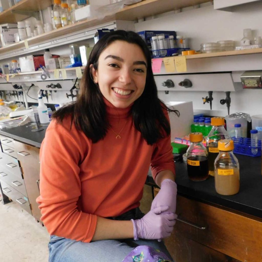Marya Ornelas '20 sits in a science lab and wears an orange shirt and light purple gloves.