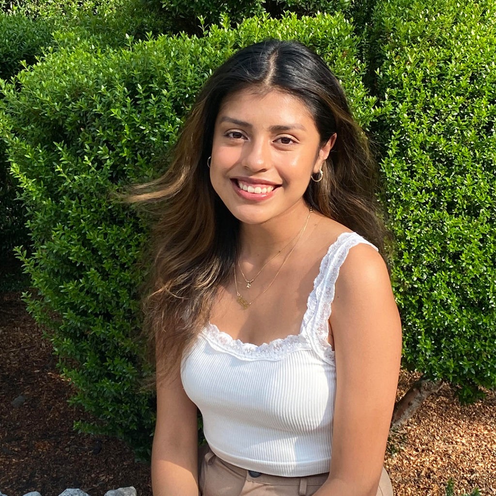 Xitlaly Franquez '23 has long brown hair and wears a white stop while sitting in front of a green bush.
