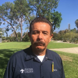 Carlos Ortega stands on the Mounds with trees in the backdrop and wears a dark blue uniform with the Pitzer logo and his first name in white. Carlos has dark brown hair and mustache/