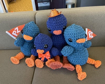 Four crocheted Cecil the Sagehens sit on a couch. They range from dark to light blue and have large round black eyes and orange bills and feet. One wears an orange bowtie and one wears a glittery orange hat. The ones on the right and left hold up orange felt banners with PZ in white letters.