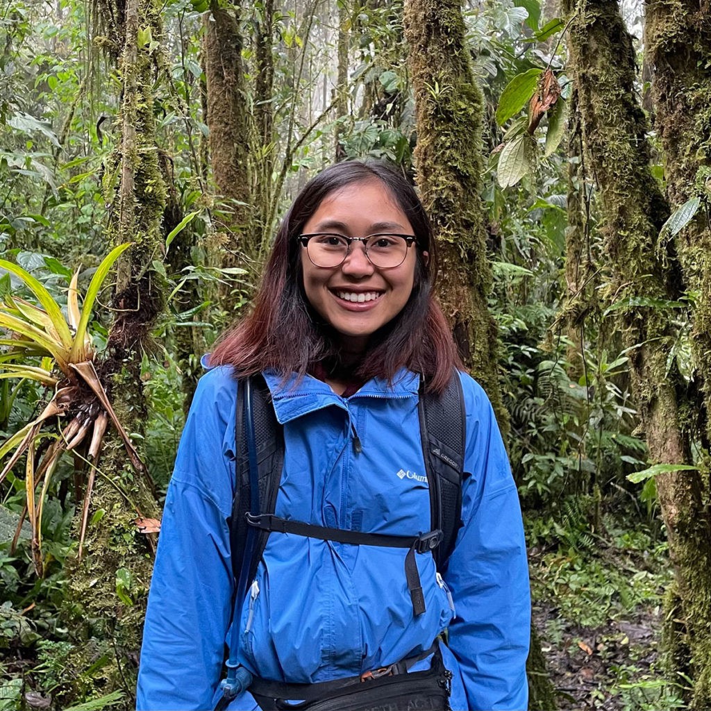 Chanchanok Sudta wears a backpack and blue jacket as she stands in a lush green forest and smiles at the camera.