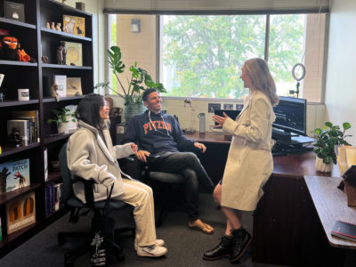 Huei Ming Lim and Professor Marcus Rodriguez sit in rolling chairs in Rodriguez’s office and smile at Sarah Mann as she stands and leans against a desk as she raises a hand mid-conversation. Rodriguez wears a dark blue and orange Pitzer sweatshirt. A tall shelf with various children’s books and figurines are to the left and a window with a view of the trees on campus are behind them.