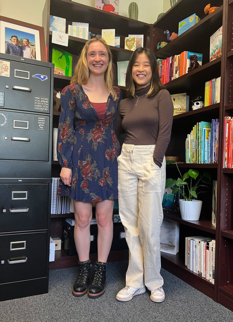 Sarah Mann and Huei Ming Lim stand side-by-side in front of a tall shelf full of bright colored children’s books and thank you cards. Mann has long straight blond hair and wears a blue dress with a red flower pattern and Lim has shoulder-length straight brown air with blond tips and wears a long-sleeved brown shirt and white pants.