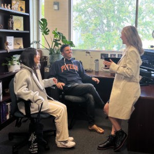 Huei Ming Lim and Professor Marcus Rodriguez sit in rolling chairs in Rodriguez’s office and smile at Sarah Mann as she stands and leans against a desk as she raises a hand mid-conversation. Rodriguez wears a dark blue and orange Pitzer sweatshirt. A tall shelf with various children’s books and figurines are to the left and a window with a view of the trees on campus are behind them.