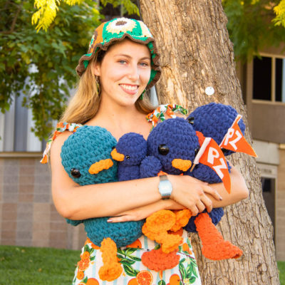 Lucy Conover has long blond hair and wears a crocheted green, orange, and brown cap and an outfit with light blue stripes and a pattern of oranges. She holds four crocheted stuffed blue Cecil sagehens with orange bills and feet and large round black eyes. Two Cecils hold triangle orange flags with PZ in white letters.