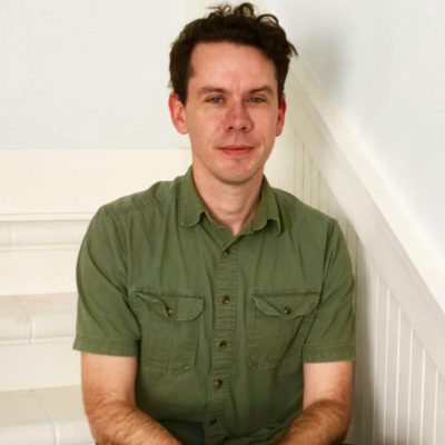 Quentin Ring has short dark brown hair and wears an olive green collared shirt as he sits on a white staircase.