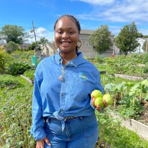 Yaquana Williams '21 in a blue collared shirt with the FoodCorps logo stands in a green garden and holds three bright green round vegetables.