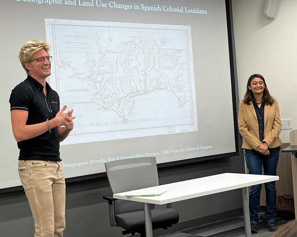 Ben Willett and Urmi Engineer Willoughby stand on the left and right side of a projector screen with  a line drawing map of Louisiana with the title Demographic and Land Use Changes in Spanish Colonial Louisiana at the top.