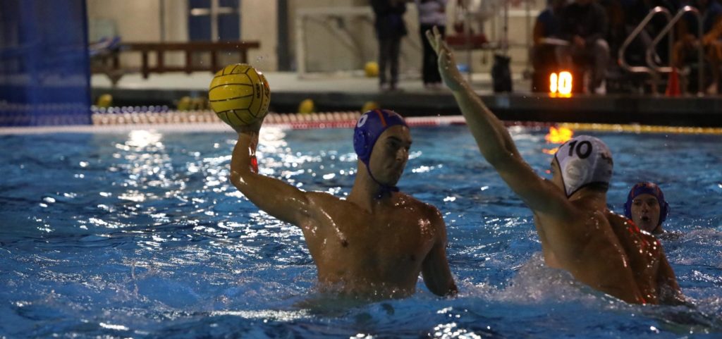 A player from the Pomona-Pitzer men’s water polo team wears a blue swim cap and holds up a yellow ball as an opposing player in a white swim cap raises an arm to try to block it. They are in the pool at nighttime.