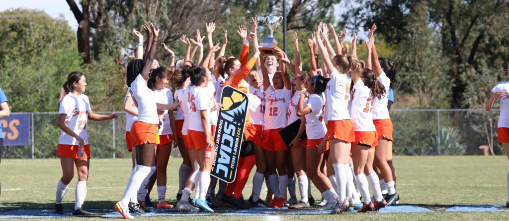 Members of the Sagehen women’s soccer team in their orange and white uniforms raise their hands in celebration and crowd around a teammate holding up a silver cup trophy. A sign reading 2022 Women’s Soccer Tournament Champions dangles from another teammate’s hand.
