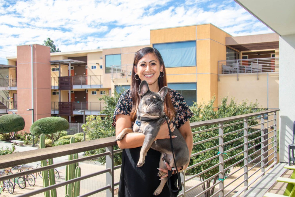 Steffanie Guillermo stands on her balcony with a backdrop of cacti and the orange and beige residence halls under a cloudy blue sky. She holds her gray French bulldog Solo.