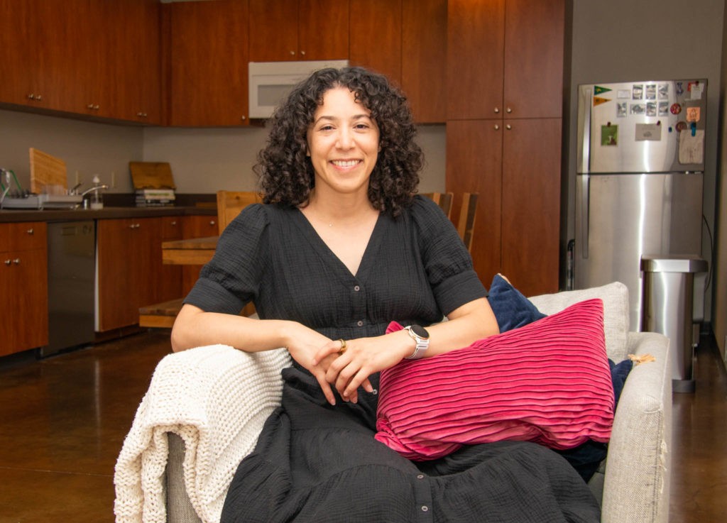 Jessica Kizer sits in her apartment on a gray chair with a magenta pillow on her lap. Her kitchen with sleek dark wood cabinets and a silver refrigerator are in the background.