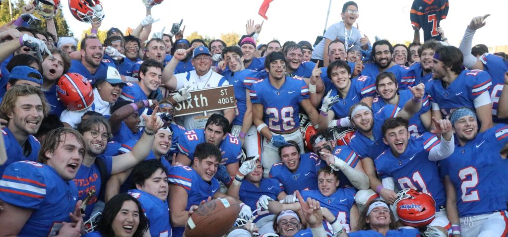 The Pomona-Pitzer men’s football team in their blue, orange, and white uniforms gather around their coach in white and blue who hold up the trophy with the brown Sixth Street sign on top.