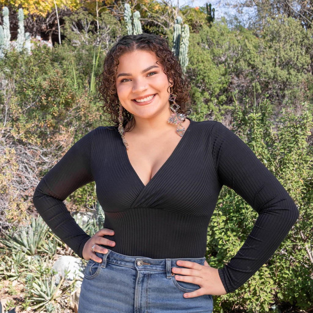 Jansikwe Medina-Teyac has shoulder-length curly brown hair and wears a long-sleeved V-neck black shirt and blue jeans. She puts her hands on her hips and stands in front of a backdrop of Pitzer’s native plants of cacti, trees, and succulents.