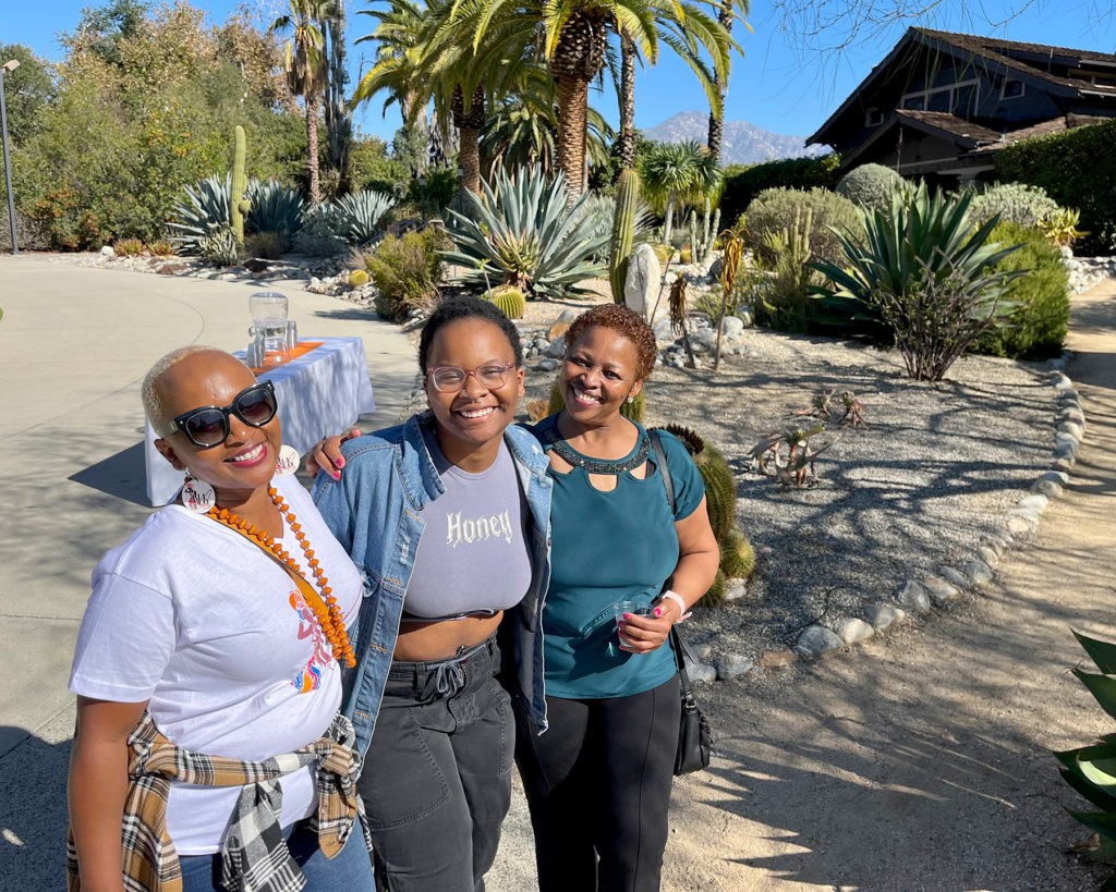 Lillian Latan and Joyce Ngatia put their arms around Lillian’s daughter Lola as they smile and stand in front of the Grove House with palm trees and desert landscaping in the background.
