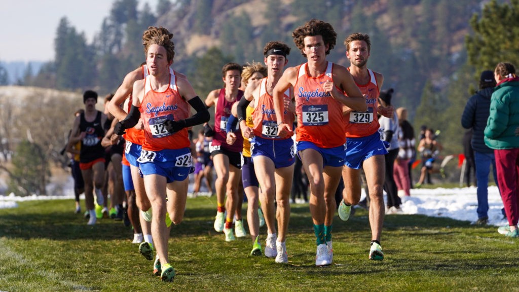 The Pomona-Pitzer men’s cross-country team are wearing their uniform of orange Sagehens shirts with blue shorts as they run across a green field with trees and mountains in the backdrop.