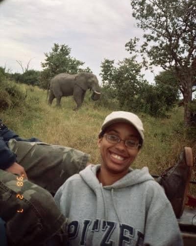 Sayjal Waddy is pictured as a student on a safari with an elephant walking among the grass and trees in the background. Waddy ears a white baseball cap and gray sweatshirt.