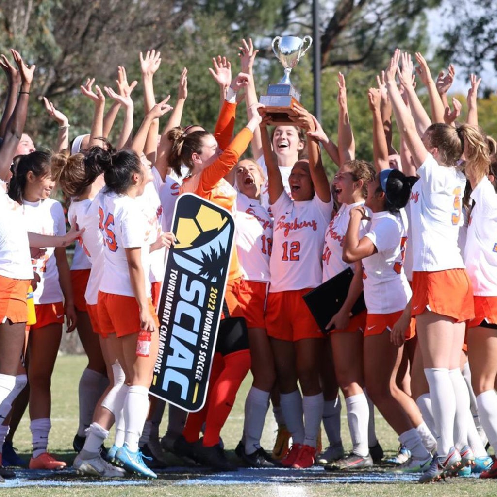 Members of the Sagehen women’s soccer team in their orange and white uniforms raise their hands in celebration and crowd around a teammate holding up a silver cup trophy. A sign reading 2022 Women’s Soccer Tournament Champions dangles from another teammate’s hand.