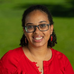 Sayjal Waddy wears a red shirt, red rectangular dangling earrings, and black-rimmed glasses. She has shoulder-length curly black hair that is tied back. The background is a blurred green lawn.