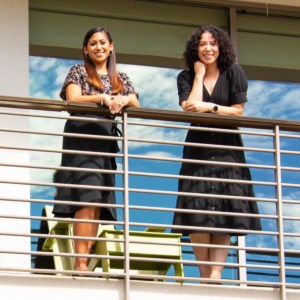 Steffanie Guillermo and Jesica Kizer lean against the railing on Guillermo’s second-floor balcony. Steffanie Guillermo has long straight dark brown hair with auburn tips and wears hoop earrings, a floral shirt, and long black skirt. Jessica Kizer has shoulder-length curly black hair and wears a watch and a long black dress with white buttons.