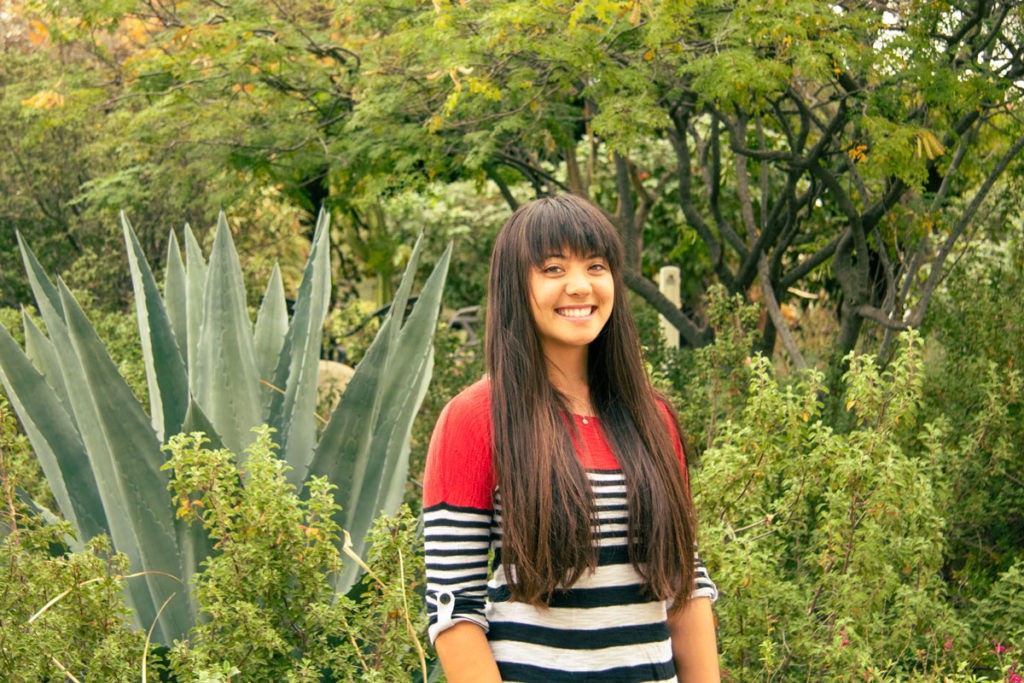 Shelby Ottengheime stands against a backdrop of green trees and succulents. She wears a long-sleeved shirt with red at the top and black and white stripes at the bottom. She has long straight brown hair and bangs.