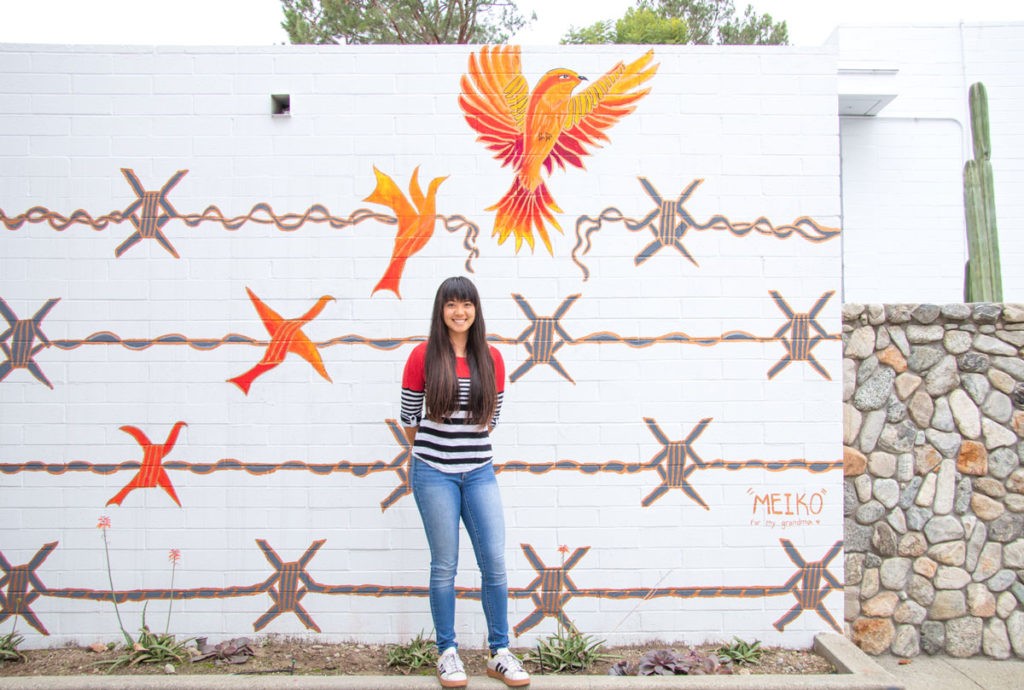 Shelby Ottengheime stands in front of the white Mead Hall wall with her mural, which depicts gray barbed wire with a barb that transforms into a bright orange and yellow bird that opens its wings in flight at the top right corner. In the bottom right corner text reads “‘Meiko’ for my grandma.” Shelby wears blue jeans and a long-sleeved shirt with red at the top and black and white stripes at the bottom. She has long straight brown hair and bangs.