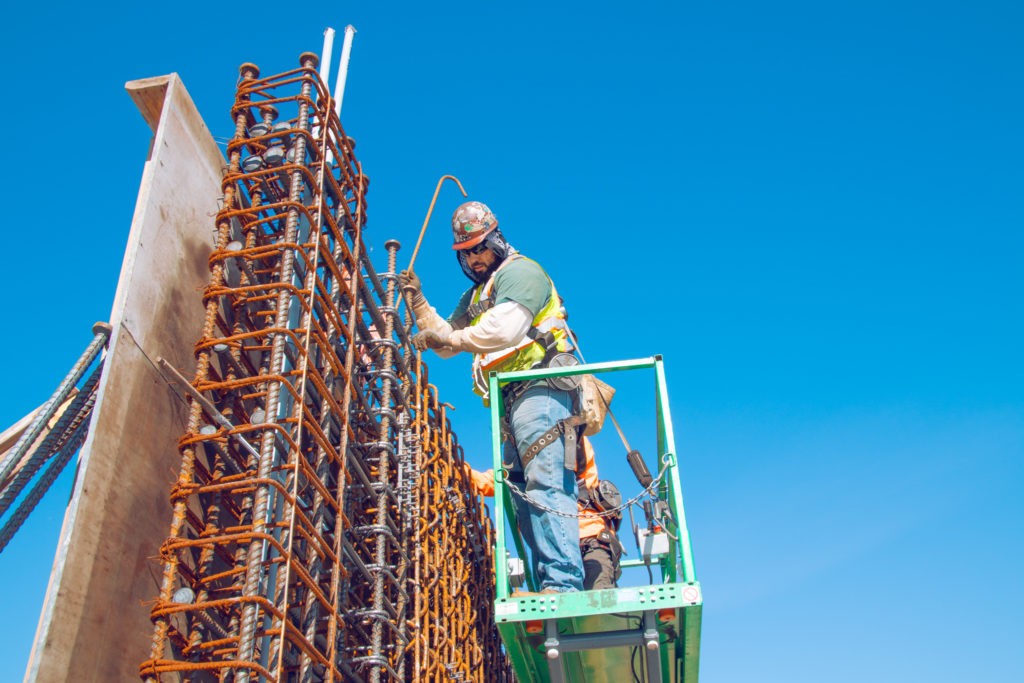 A construction worker in an orange hard hat stands in a green lift while working on a rebar column.
