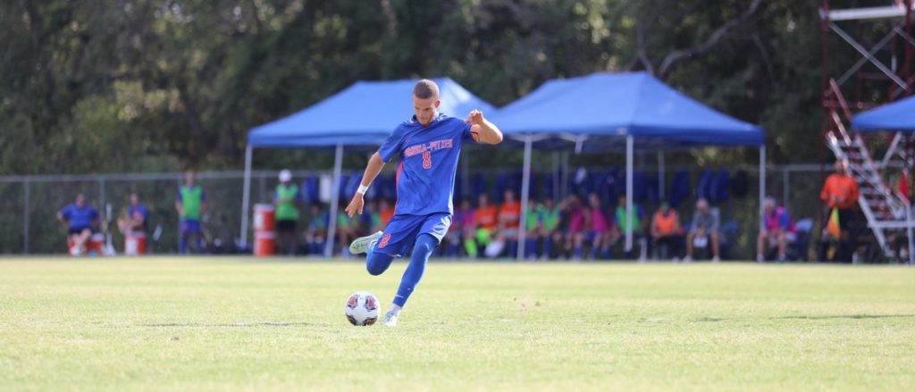 Matthew Carlson wears a blue soccer uniform with orange text on the front of his shirt that reads Pomona-Pitzer 8. He is about to kick a soccer ball on a pale green field.
