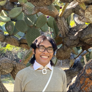 Dominic Arzadon has short dark brown hair and wears black-rimmed glasses and a grayish green sweater. Arzadon stands in front of a cactus tree.