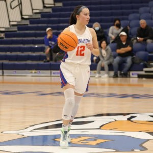 Madison Quan wears white sneakers and a white basketball uniform with blue and orange accents and the number 12 in orange on the front. Quan runs across the court while dribbling a basketball and has long dark hair pulled back in a ponytail.
