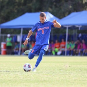 Matthew Carlson wears a blue soccer uniform with orange text on the front of his shirt that reads Pomona-Pitzer 8. He is about to kick a soccer ball on a pale green field.