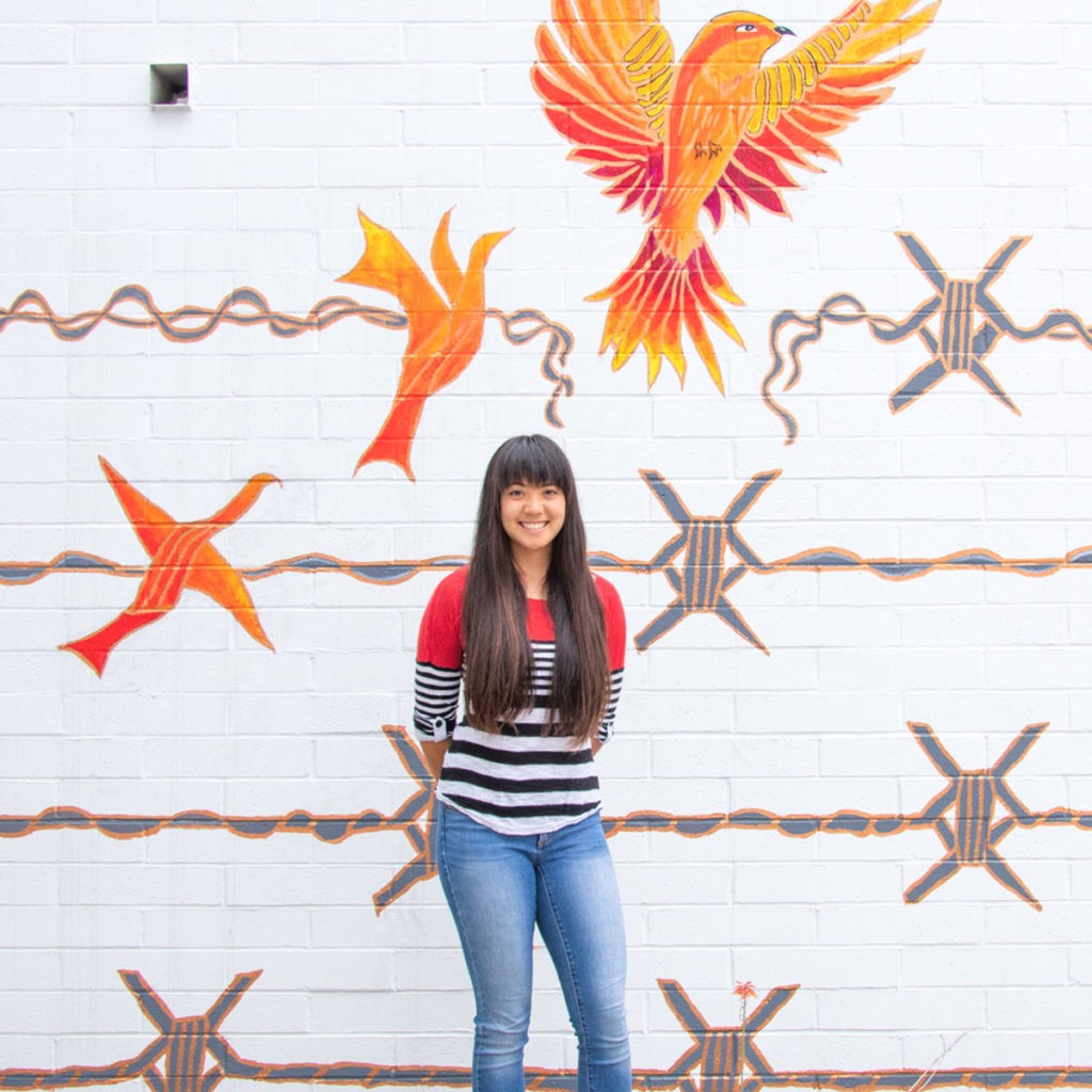 Shelby Ottengheime stands in front of the white Mead Hall wall with her mural, which depicts gray barbed wire with a barb that transforms into a bright orange and yellow bird that opens its wings in flight at the top right corner. Shelby wears blue jeans and a long-sleeved shirt with red at the top and black and white stripes at the bottom. She has long straight brown hair and bangs.