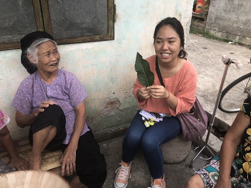 An elderly village woman and a college student sit behind a house as the student holds up a large green leaf. The student wears an orange top and blue jeans and orange shoes. The woman wears a purple shirt and black pants and hat.