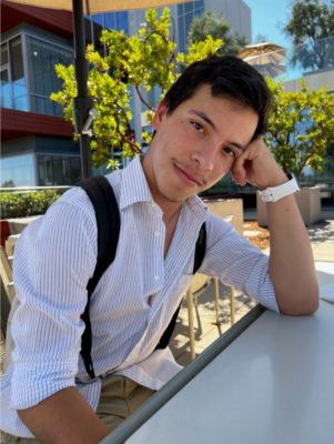 Daniel Bonilla has short dark brown hair and wears a backpack and a white collared shirt with thin blue stripes. He rests his head on his fist as he leans his elbow on a white table.