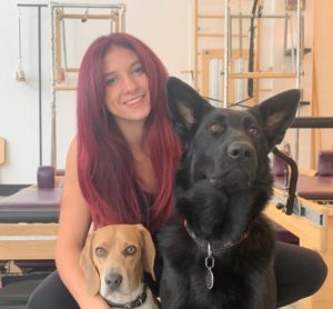 Danika Petit has long straight maroon red hair and poses with a large black dog with pointy ears and a small brown dog with floppy ears.