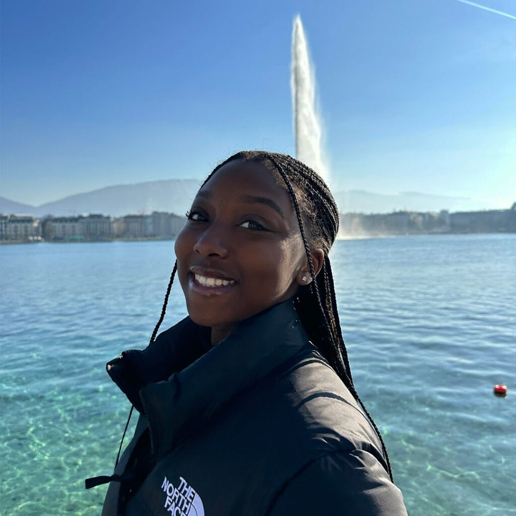 Fatoumata Dioubate has long black hair in a series of braids and wears a puffy black coat with The North Face logo. Behind her is clear blue water with a fall foundation of water bursting up.