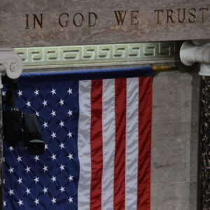 The American flag hangs under an inscription "In God we trust" on a gray wall.