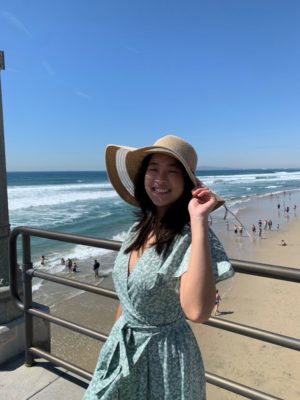 Huei Ming Lim has long straight black hair and wears a pale blue wrap dress and a tan floppy hat. Lim leans against a black railing with a beach in the background.