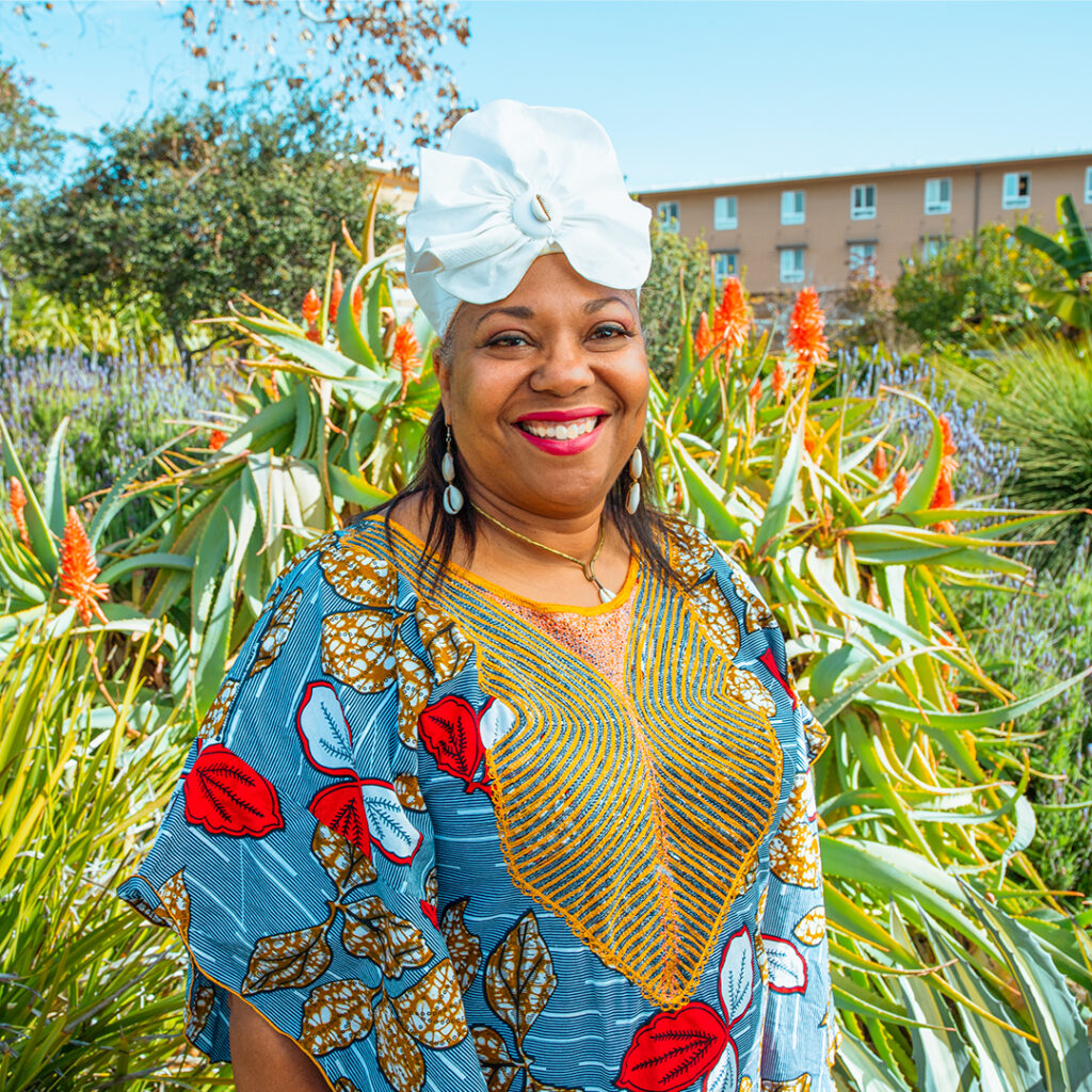 Leeshawn Cradoc Moore wears a West African light blue dress with prints of red/light blue and dark gold/light blue leaves, a white hat with a seashell in the center, and white seashell earrings. The campus greenery and a dorm is in the backdrop.