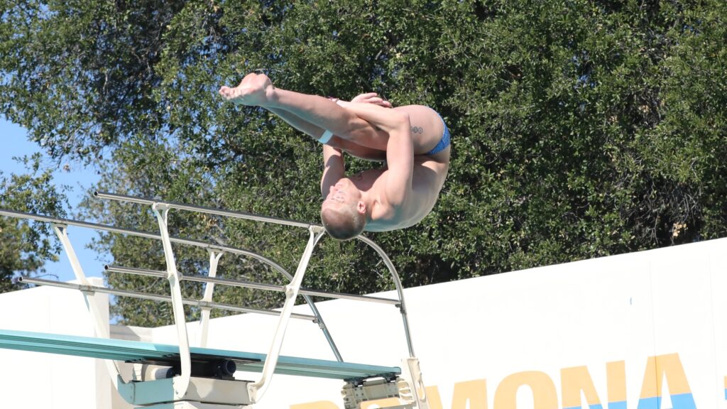 Ben Willett is upside-down in mid-flip during a dive. His legs are straight as he wraps his arms behind his knees. In the backdrop is a diving board and green tree foliage.