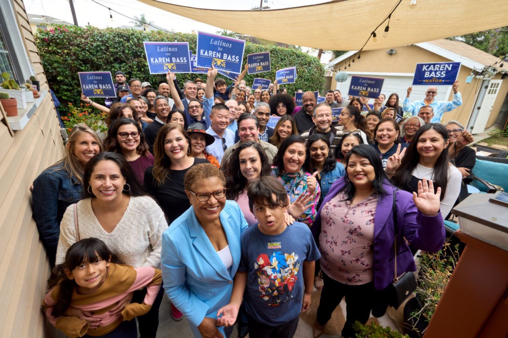 A crowd gathers around Karen Bass in the backyard of a pale yellow house. Standing in front, Bass has a short afro of brown hair and wears glasses and a sky blue blazer with white undershirt. Toward the back, a dozen people hold up navy blue signs with white text that reads Latinos for Karen Bass.
