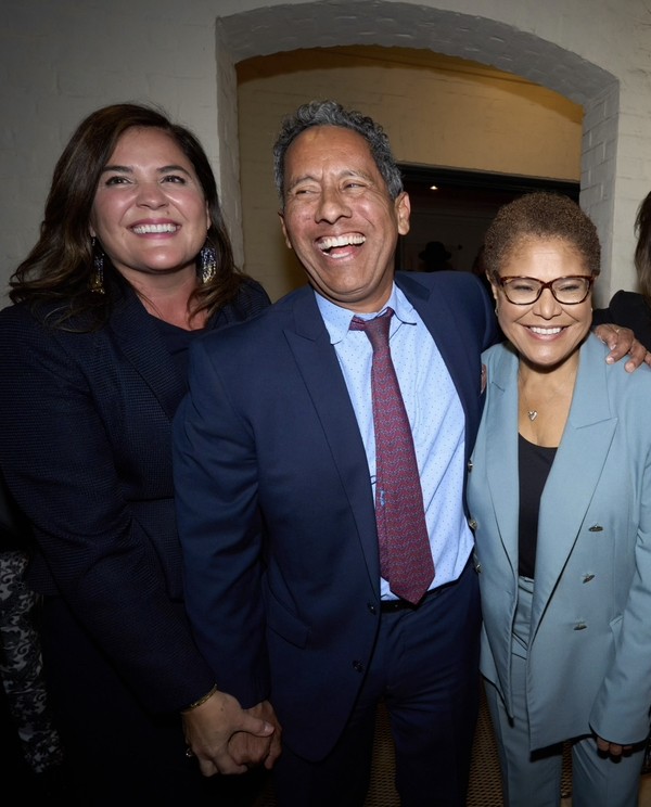 Michele Siqueiros holds her partner Solomon Rivera’s hand as Solomon puts an arm around Karen Bass. Siqueiros wears a navy blue blazer and has straight shoulder-length brown hair. Rivera has short salt and pepper hair and wears a navy blue suit with light blue dress shirt and maroon tie. Bass has a short afro of brown hair and wears glasses and a light blue blazer and pants.