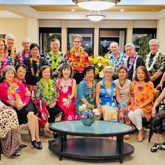 A group of Asian ballroom dancers wearing colorful dresses and Hawaiian shirts and leis. Women sit in the front row and men stand behind them.