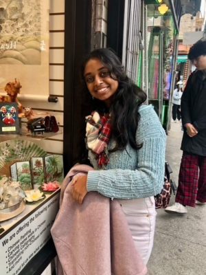 Pratya Poosala has long wavy black hair and wears a pale blue sweater, white pants, and a plaid red and white scarf. Poosala holds a pale pink coat while standing beside a store’s window display.