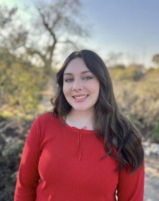 Sara Orr has long wavy brown hair and wears a long-sleeved red shirt with a red ribbon tied at the top. Behind Orr is a blurred backdrop of trees and California indigenous plants.