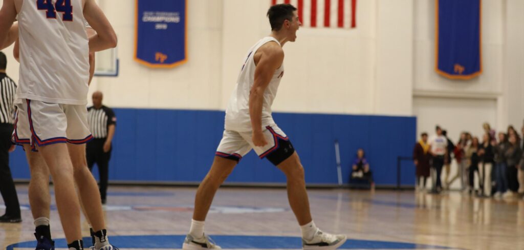 A Pomona-Pitzer basketball player strides forward, clenches his fist and shouts triumphantly. He is wearing a white uniform with blue and orange accents. The basketball court and crowd are blurred in the backdrop.