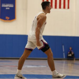A Pomona-Pitzer basketball player strides forward, clenches his fist and shouts triumphantly. He is wearing a white uniform with blue and orange accents. The basketball court and crowd are blurred in the backdrop.