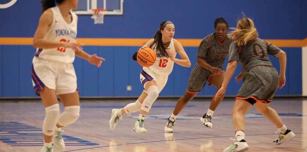 Madison Quan runs across the court while dribbling a basketball as two La Verne players attempt to block her and as Quan’s team member runs ahead. Quan has long dark hair in a ponytail and wears a blue headband and a white uniform with orange and blue accents and the number 12 in orange on the front.